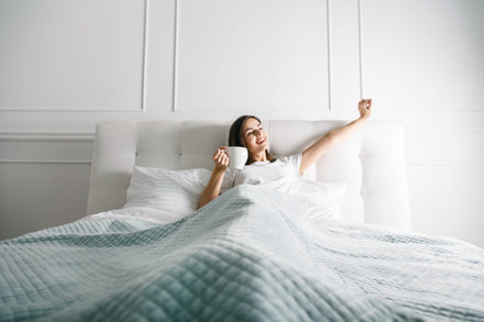 Happy woman stretching sitting on bed holding a cup of coffee