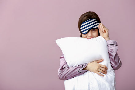 Girl standing hugging a pillow with one arm and lifting her sleep mask with the other.